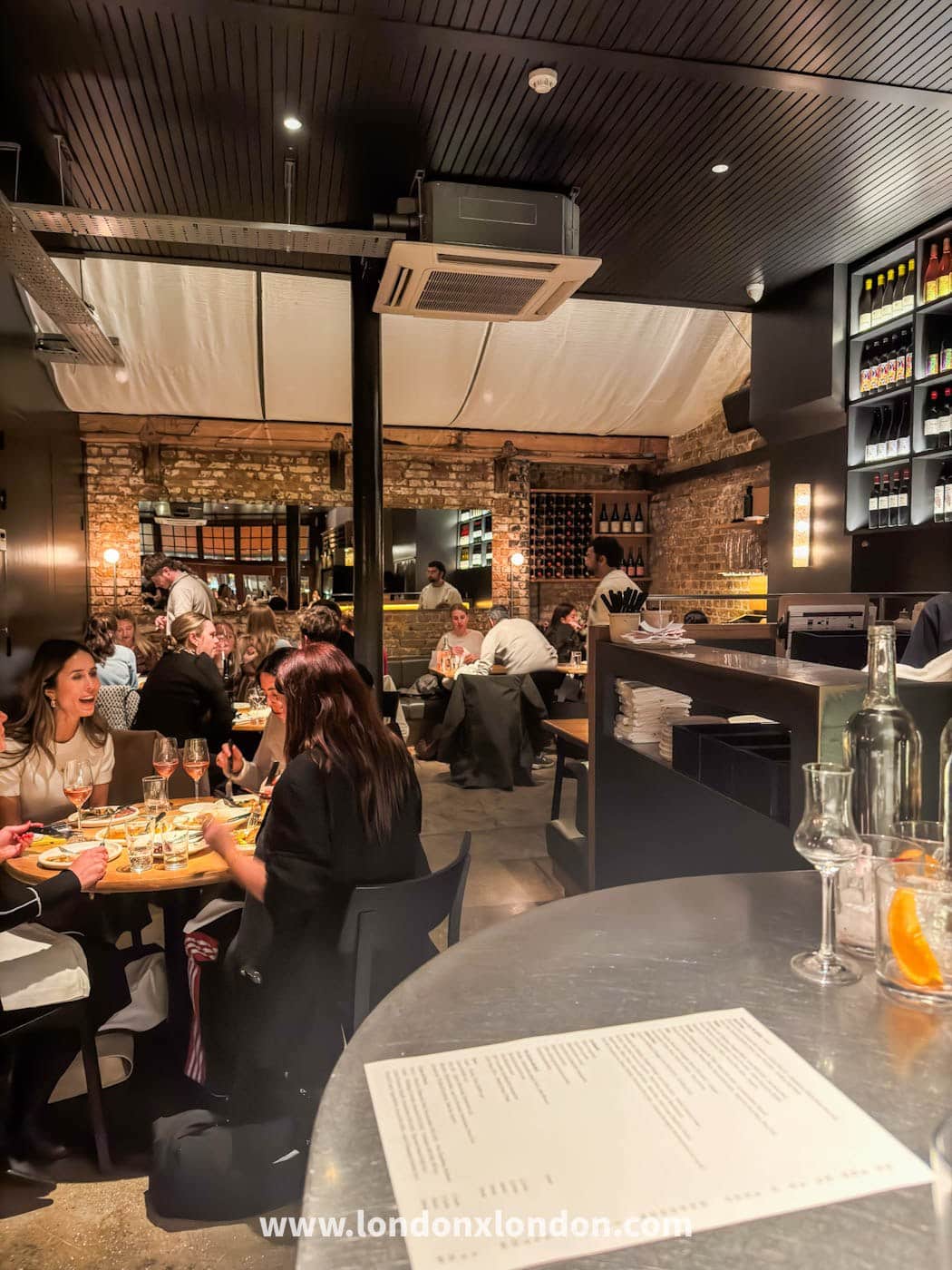 The dining room at Elliot's in Borough Market: bare brick walls, a dark slatted ceiling, zinc-topped tables of diners and a lit wall of wine bottles behind the bar