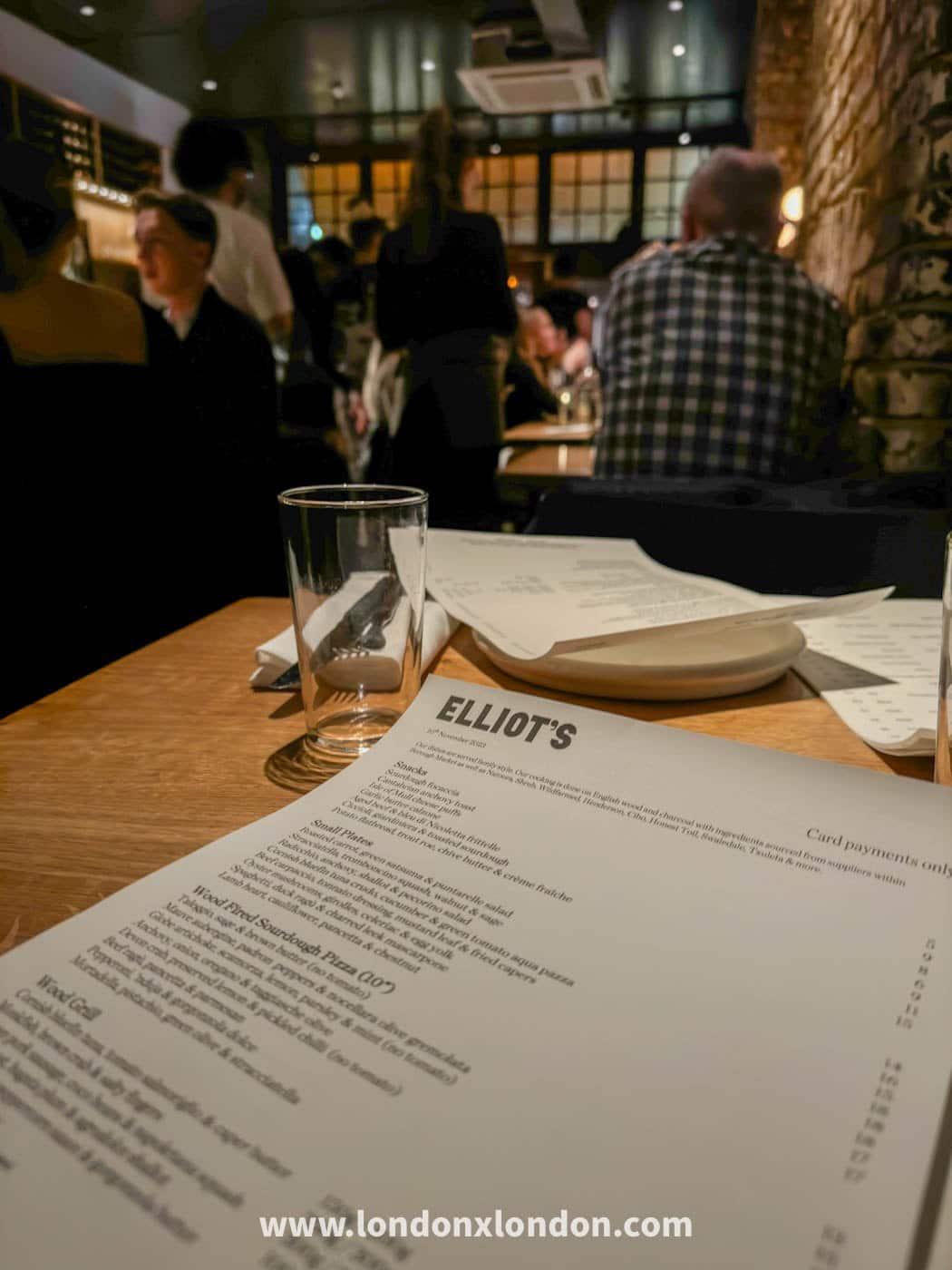 A menu on a wooden table at Elliot's, listing snacks, small plates and wood-fired sourdough pizzas, with the dining room behind