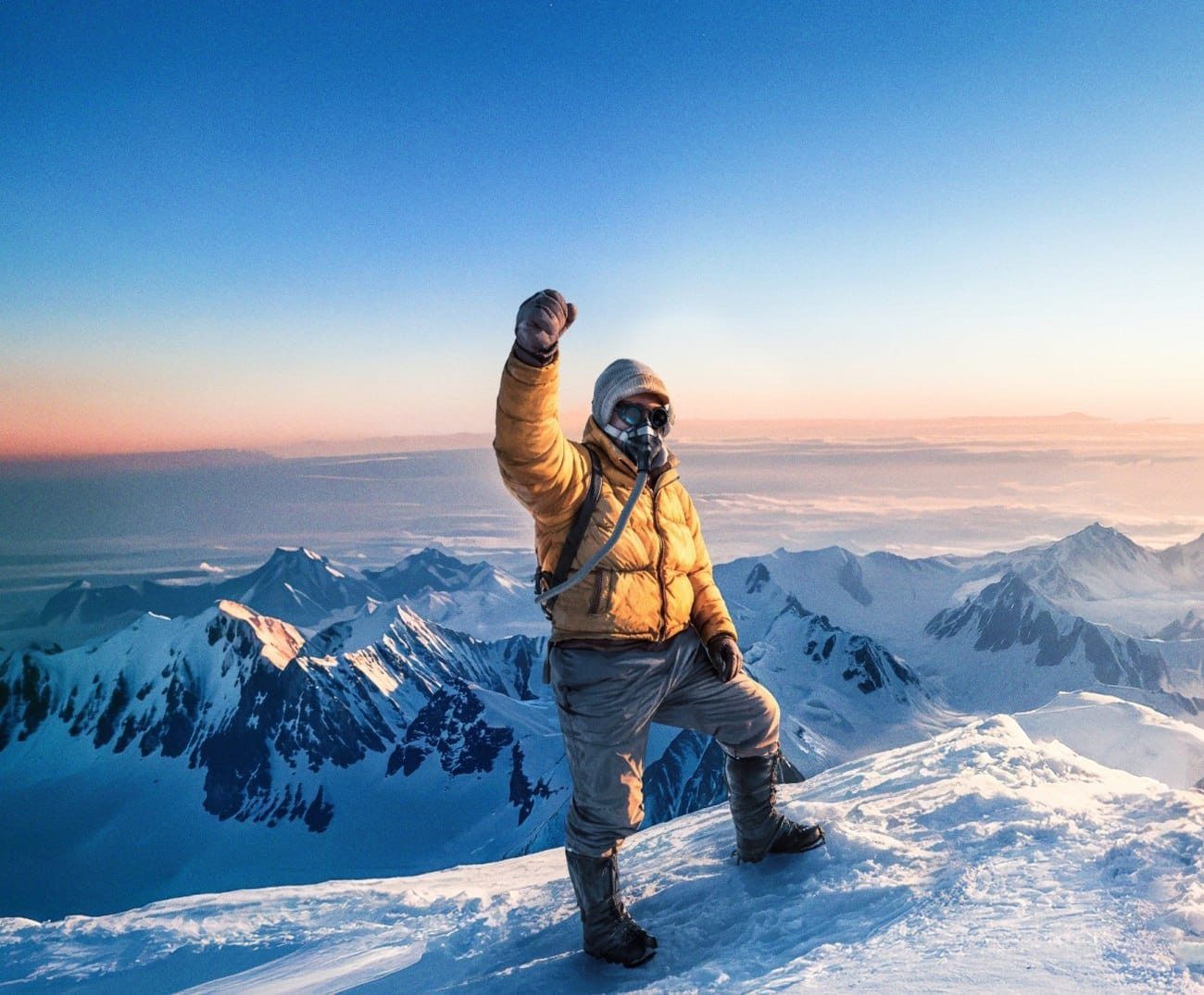 A climber in a yellow down jacket and oxygen mask raising a fist on a snow summit at dawn, with Himalayan peaks below