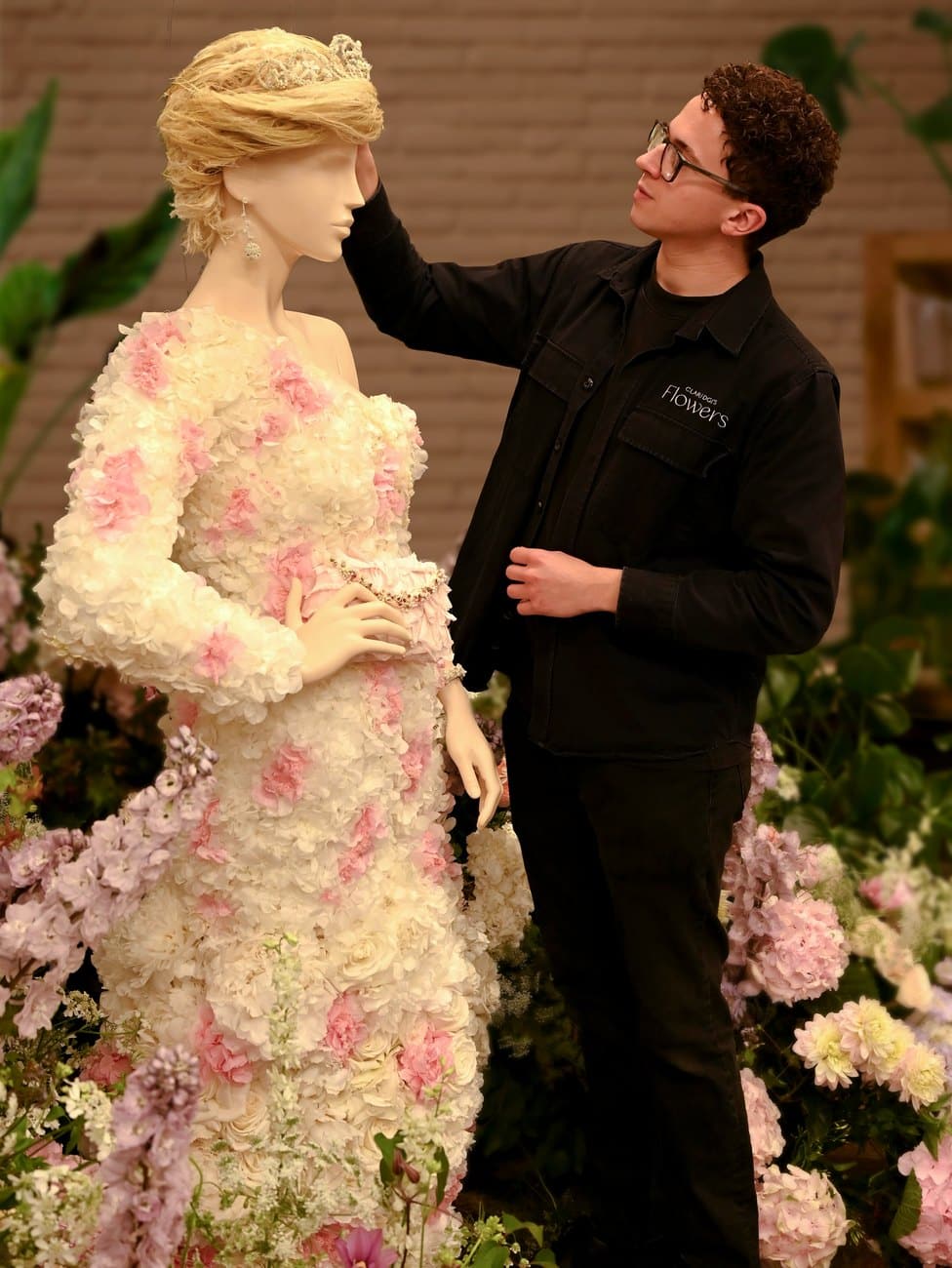 A florist in a Claridge's Flowers jacket adjusting the raffia wig on a mannequin dressed entirely in preserved white and pink hydrangea and carnation petals