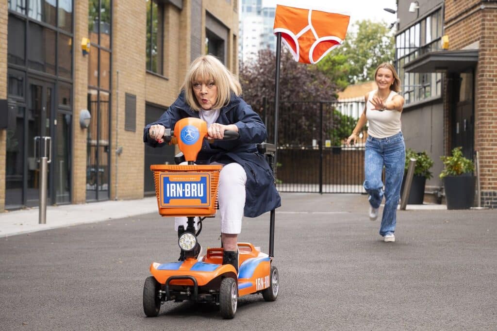 Amanda Barrie riding an orange IRN-BRU branded mobility scooter down a London street with a flag shaped like orange pants on the back, as a woman runs after her