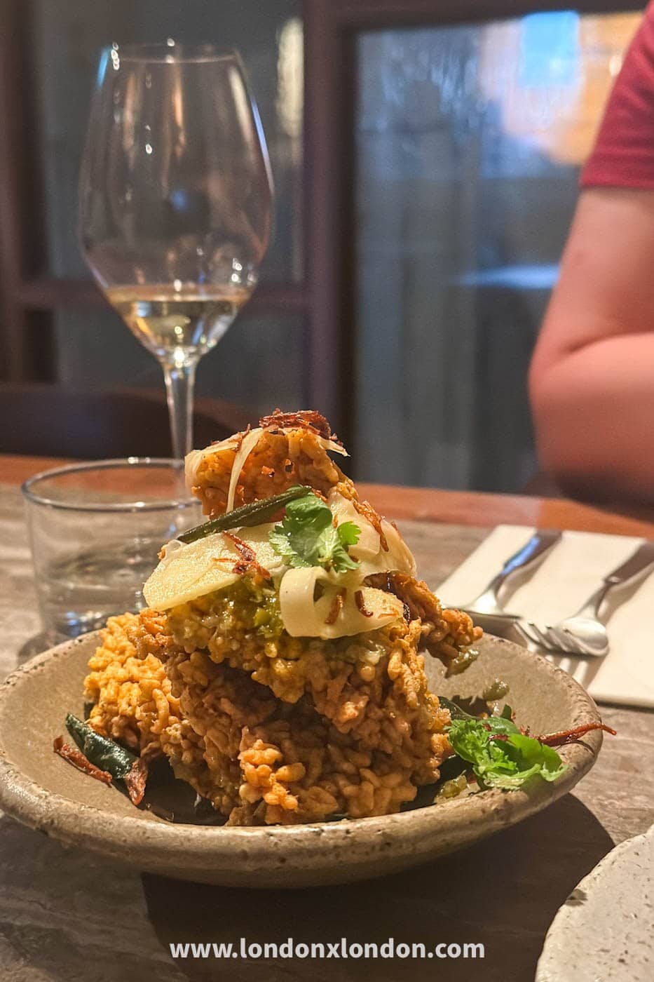 Biryani rice crackers at Kolae, stacked high in a stone bowl under crispy fried shallots, coriander and thin pale slices, with a glass of white wine behind