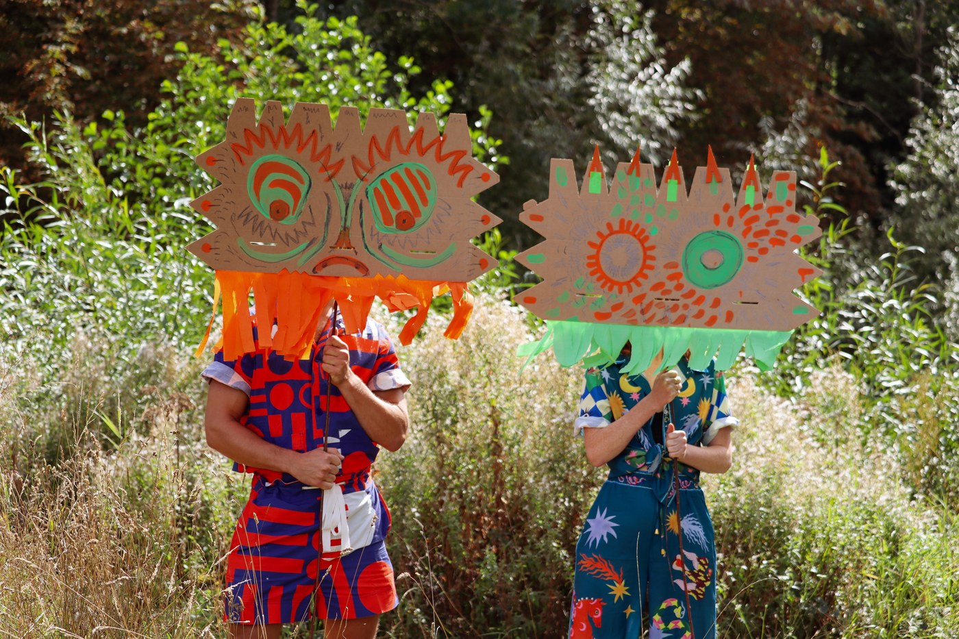Two performers in large hand-painted cardboard masks with orange and green fringing, standing in long grass