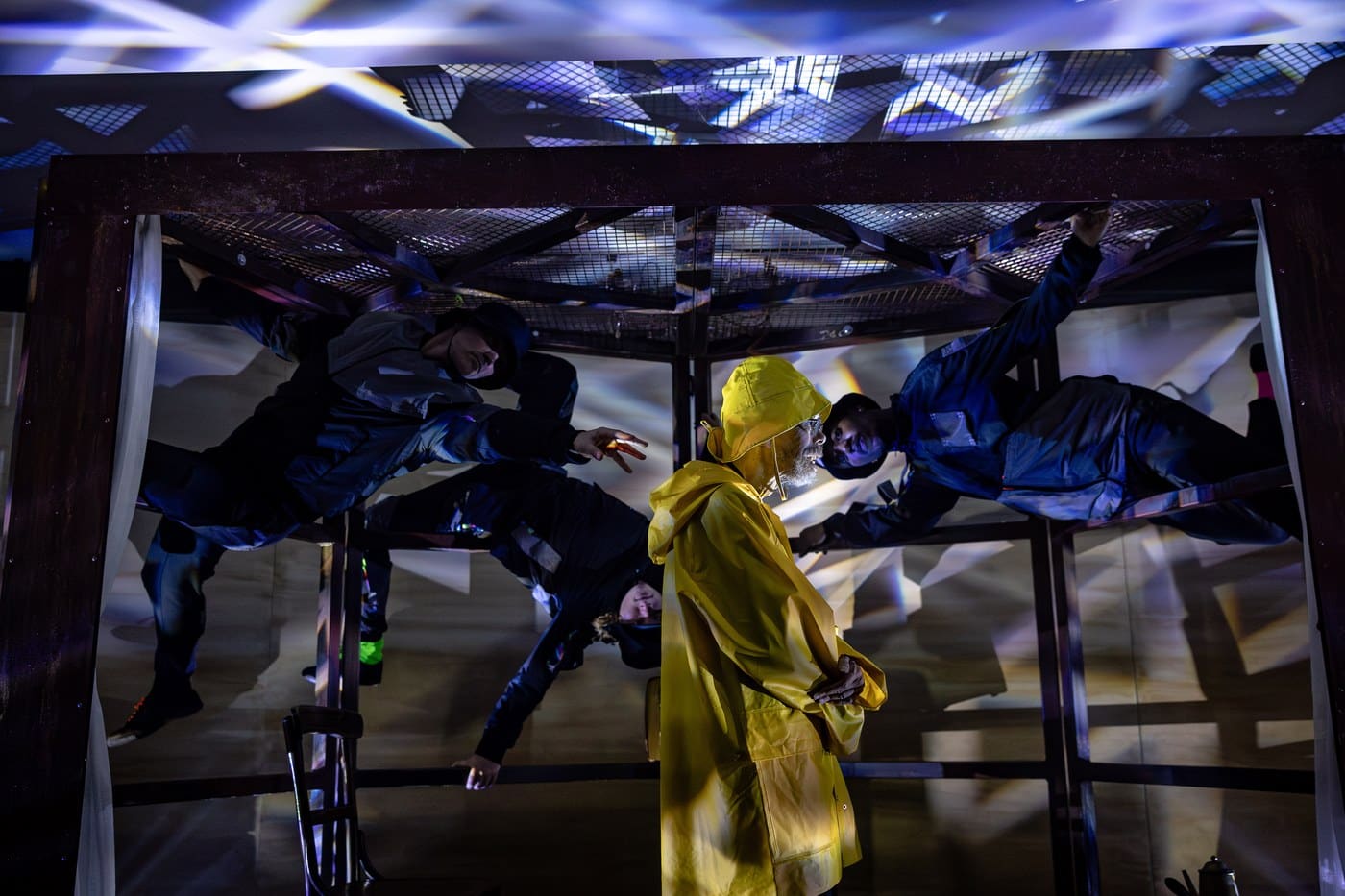 Performers suspended sideways inside a mirrored steel frame under projected light, one in a yellow raincoat standing below