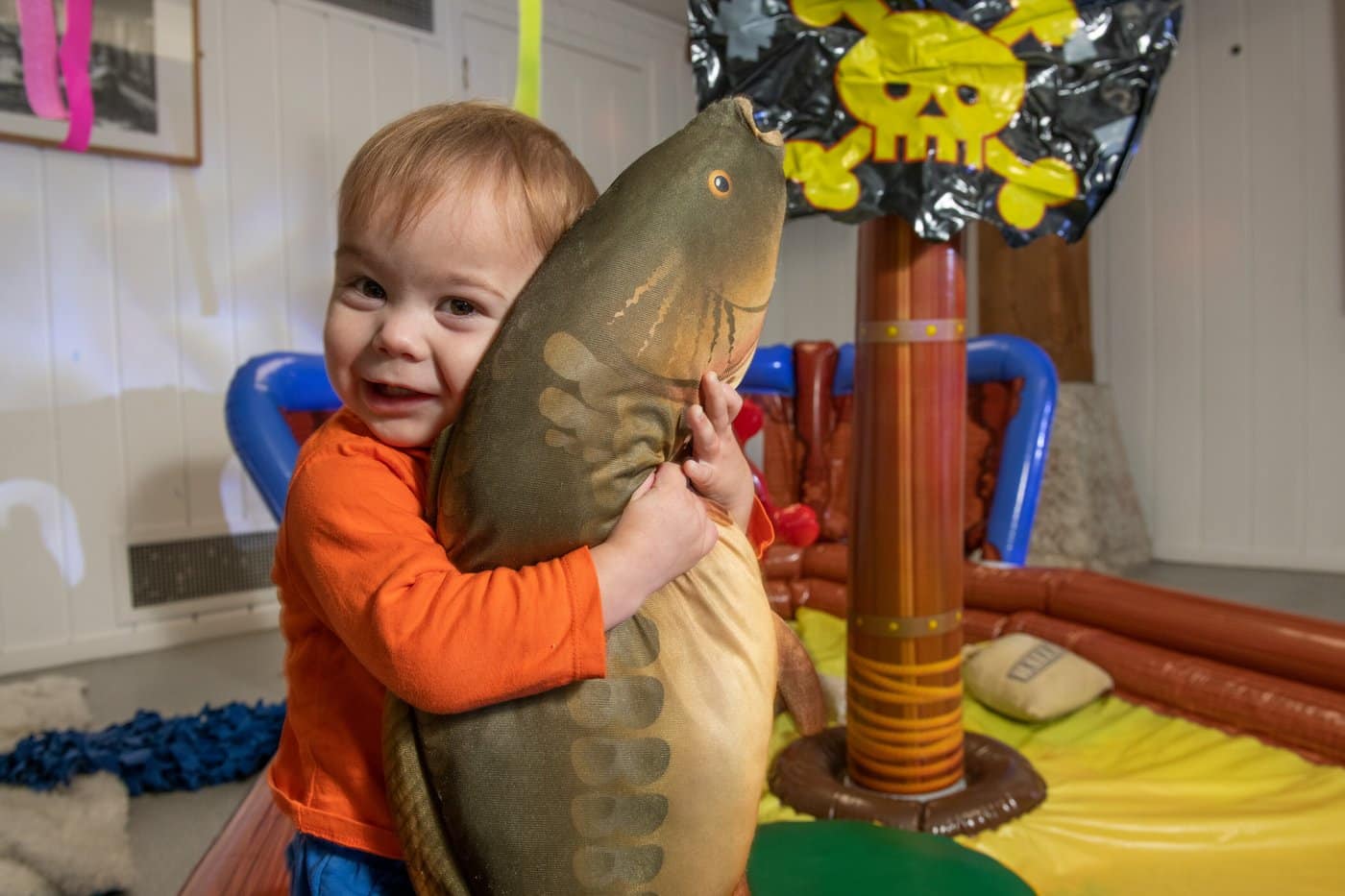 A toddler in an orange top hugging a large fish cushion beside an inflatable pirate ship at London Museum Docklands