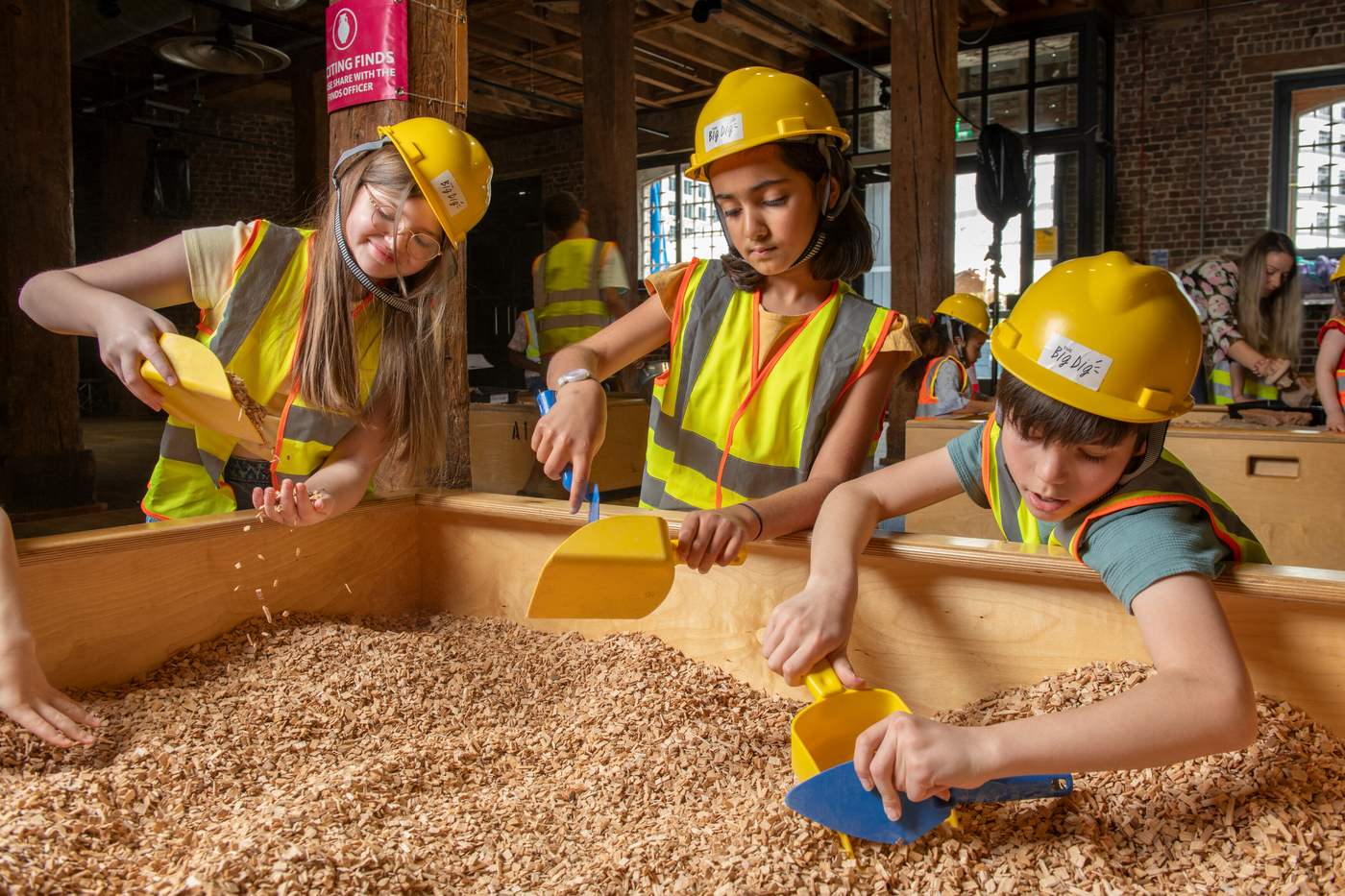 Three children in yellow hard hats and hi-vis vests digging through wood chips in a wooden trench with trowels and scoops at London Museum Docklands