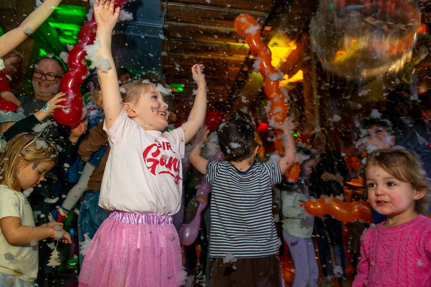 Children with hands in the air amid falling foam snow and balloons at a family rave in the warehouse at London Museum Docklands