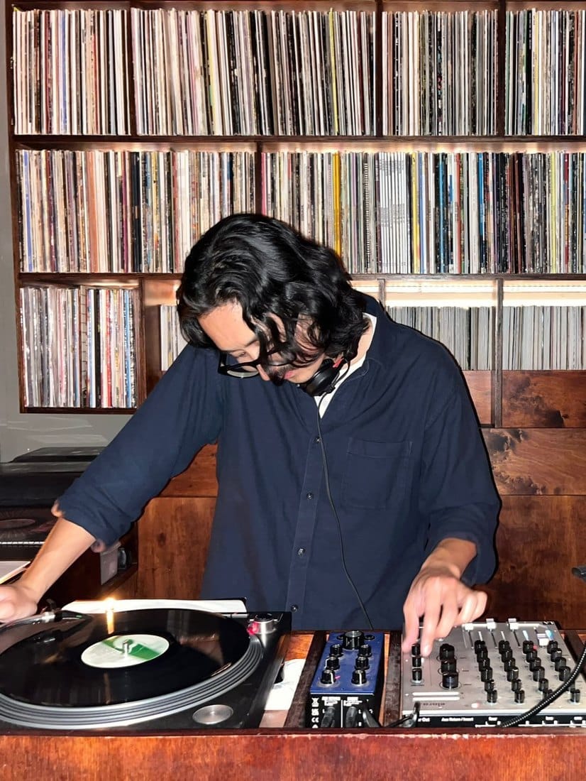 A DJ cueing a record on a turntable in front of a floor-to-ceiling wall of vinyl in wooden racks