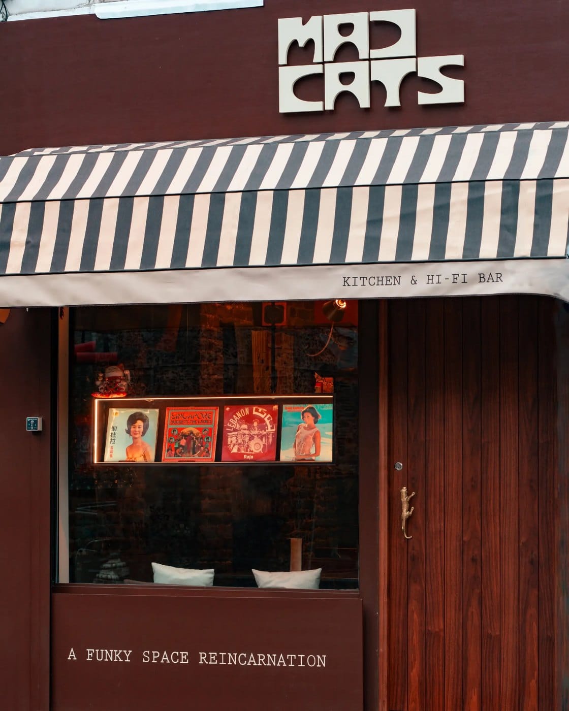 The front of Mad Cats on Redchurch Street in Shoreditch, a dark red shopfront under a black and white striped awning reading Kitchen and Hi-Fi Bar, with four record sleeves lit in the window and a brass cat door handle