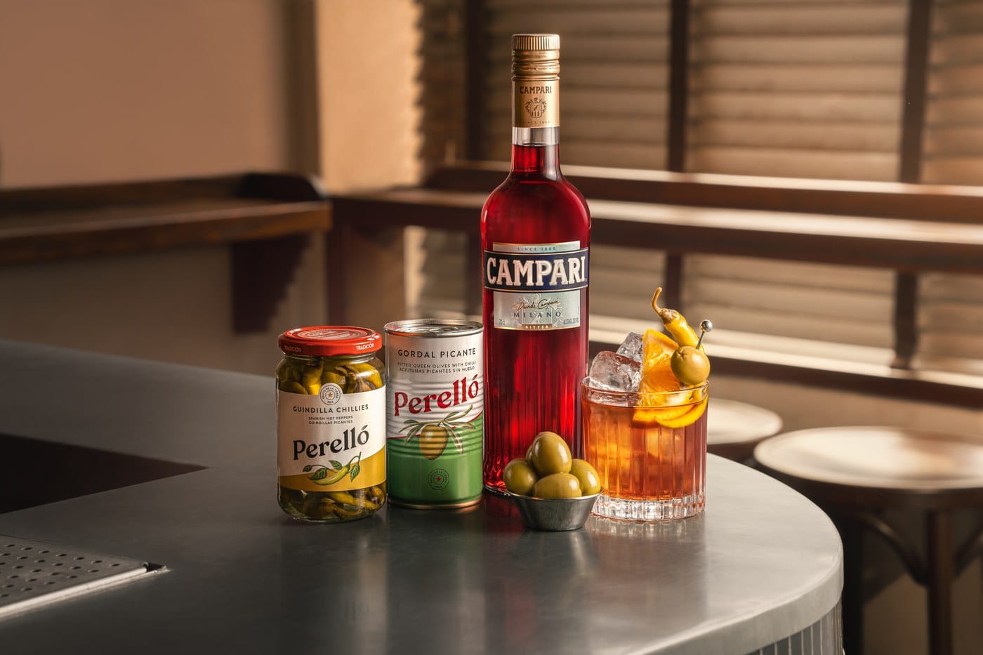 A bottle of Campari on a zinc bar top with a jar of Perello guindilla chillies, a tin of Gordal picante olives and a Negroni garnished with a chilli