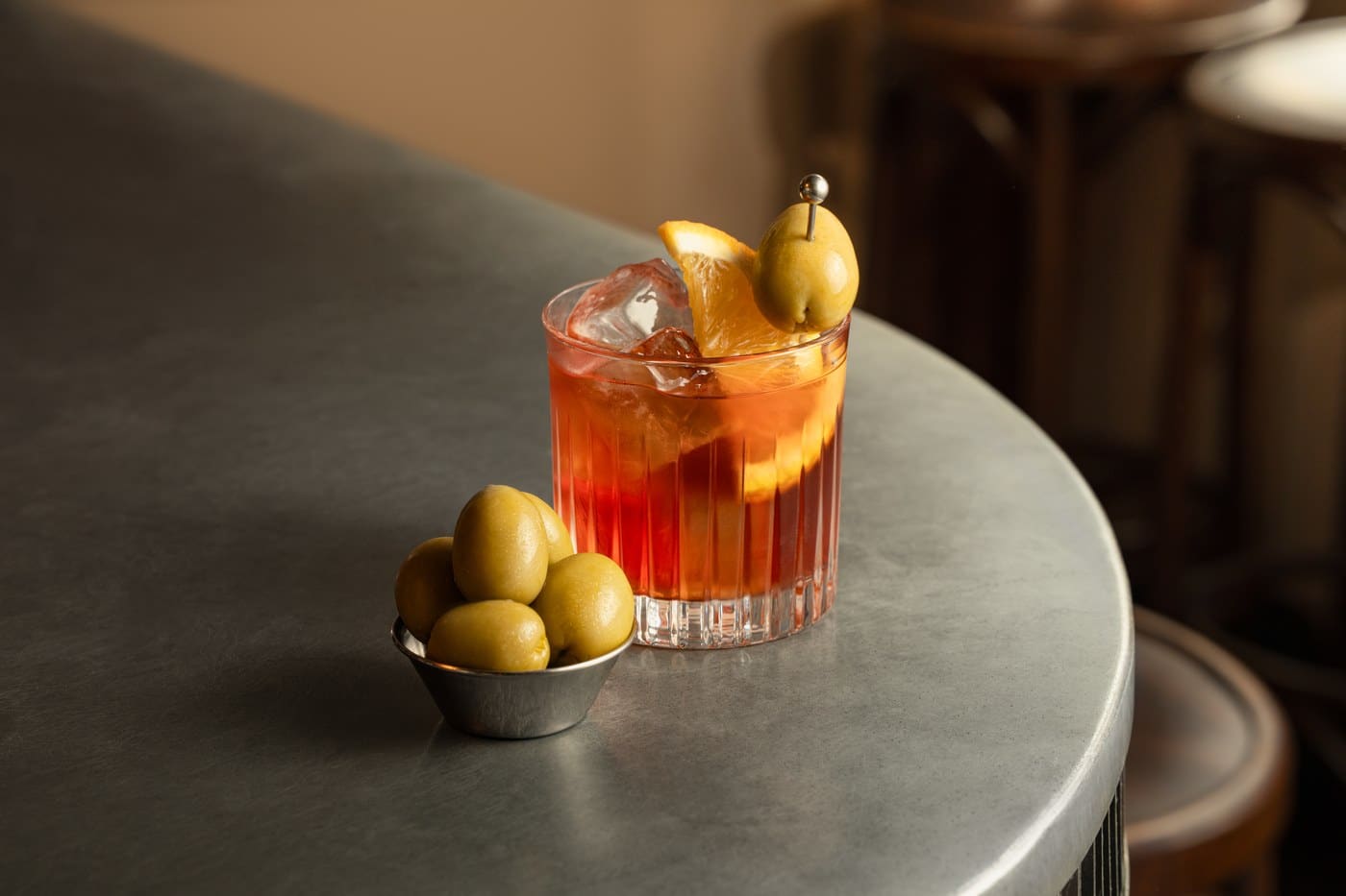 A Negroni in a ribbed tumbler garnished with an orange wedge and a green olive, beside a small metal pot of olives on a zinc bar top