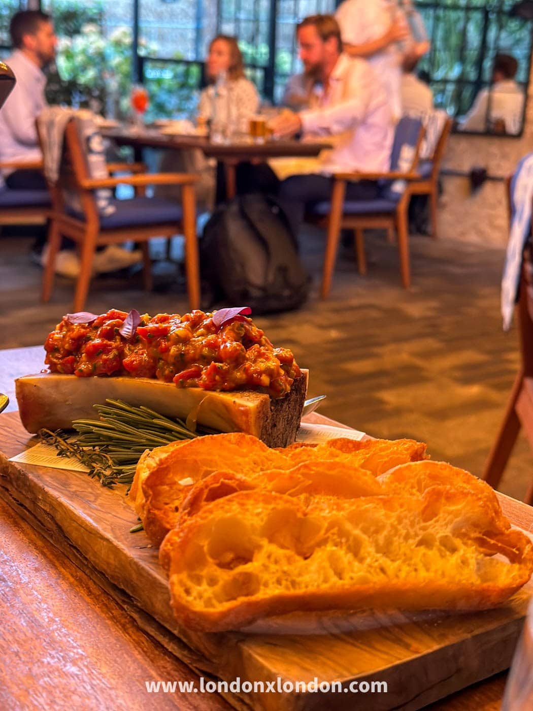 A roasted bone marrow topped with chopped tomato and pepper salsa, served on a wooden board with rosemary and thick slices of toasted sourdough at Parrillan in Borough Yards