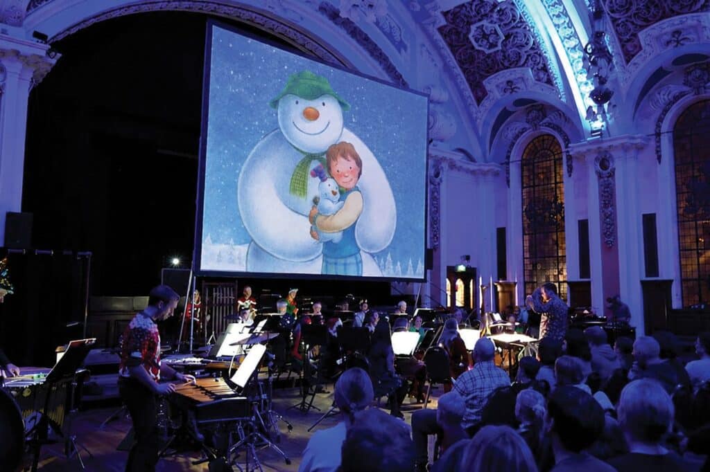 A live orchestra playing beneath a large screen showing The Snowman, in the ornate domed hall at Battersea Arts Centre