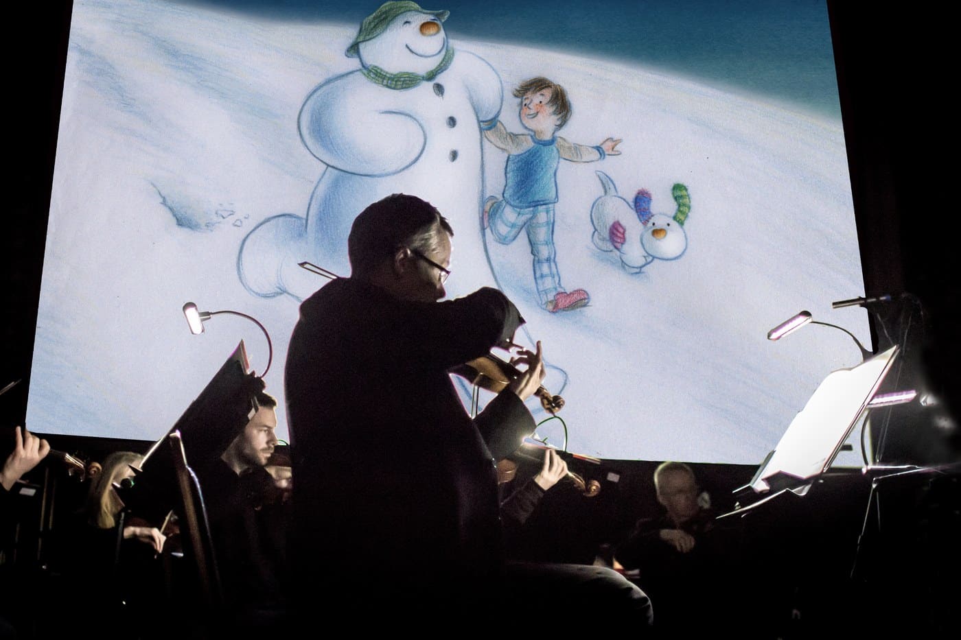 A violinist silhouetted against a screen showing the Snowman, the boy and the Snowdog in the snow