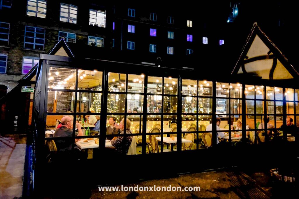 Pique-Nique restaurant lit up at night, a black-framed glass pavilion with mock-Tudor gables in Tanner Street Park, festoon lights inside and blackboard menus hung against the glass