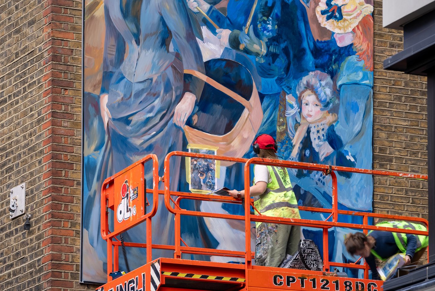 Two Global Street Art painters on an orange cherry picker working on the Renoir mural, with a small printed reference photograph of the painting taped to the wall beside them