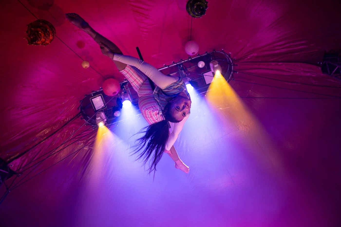 An aerialist upside down in a strap under the pink and purple canopy of a circus tent, paper lanterns hanging around her
