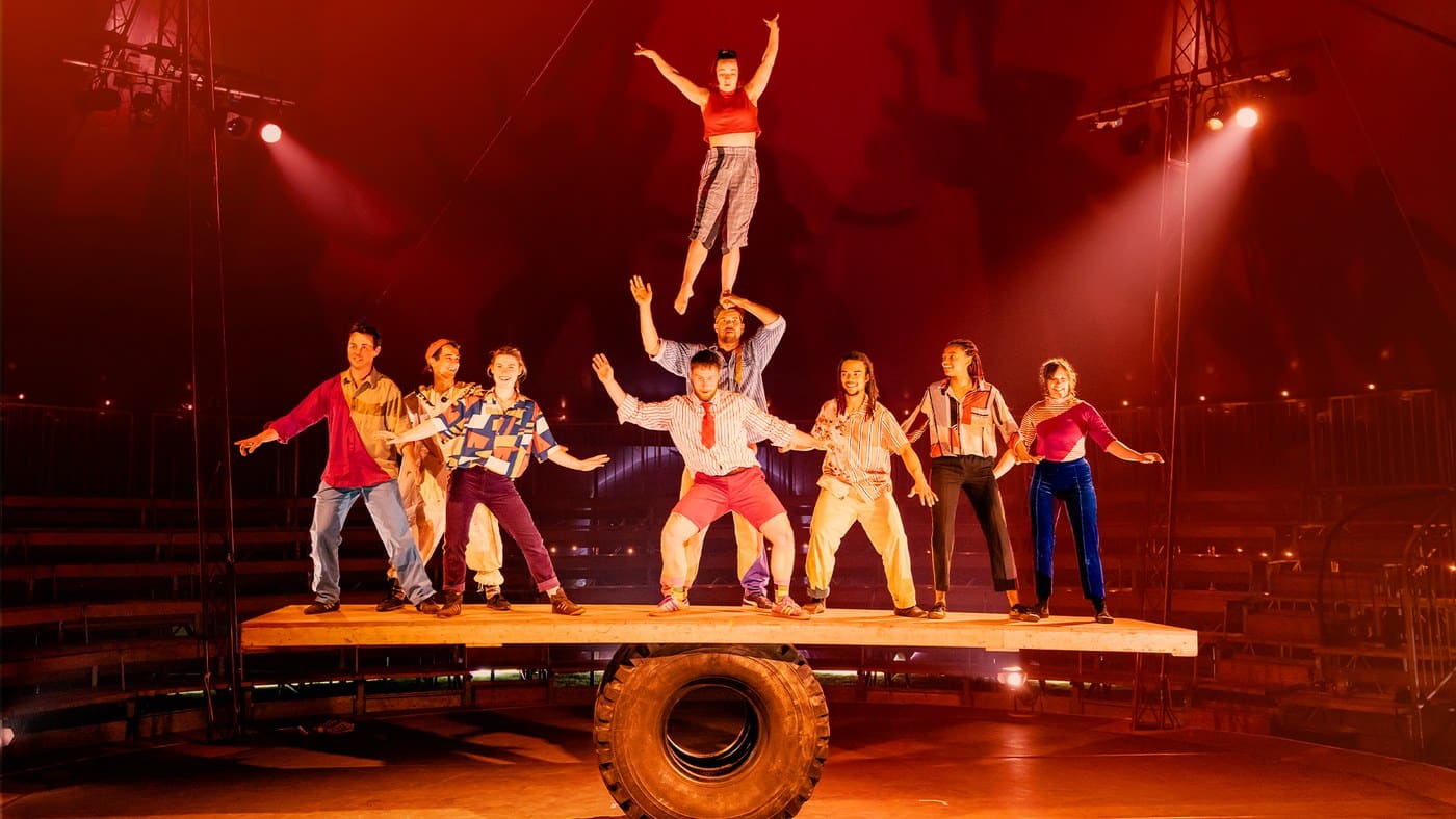 Nine performers balanced along a wooden plank resting on a tractor tyre, one standing on another's hands above them