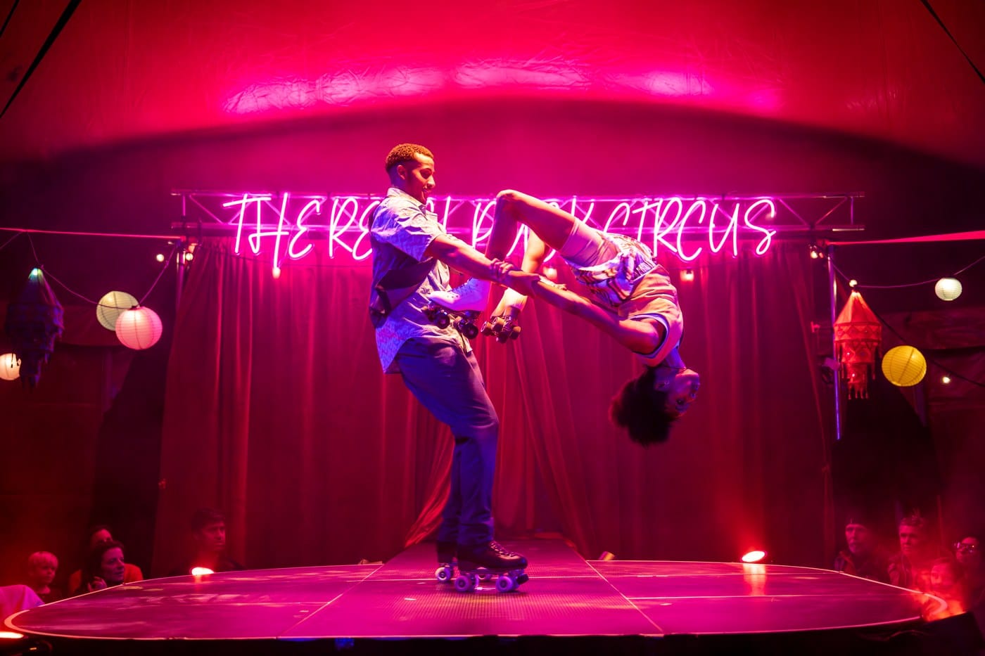 Two performers on roller skates, one holding the other upside down by the hands, beneath a pink neon circus sign