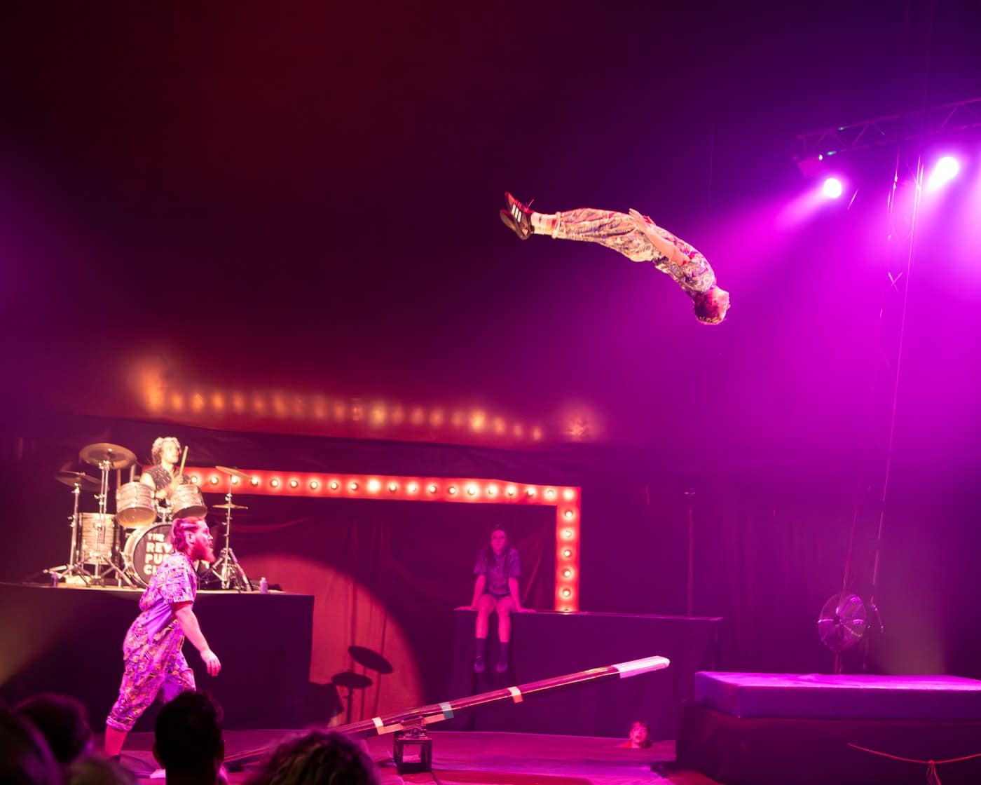 A performer mid-somersault high above a teeterboard, with a drummer and two other performers watching from below