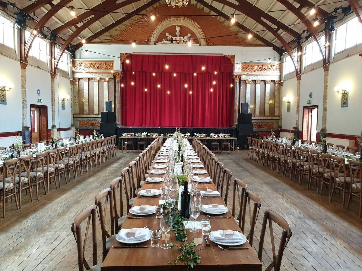 The main hall at Stanley Arts set for a long-table dinner, with a red stage curtain, festoon lights and an arched timber roof