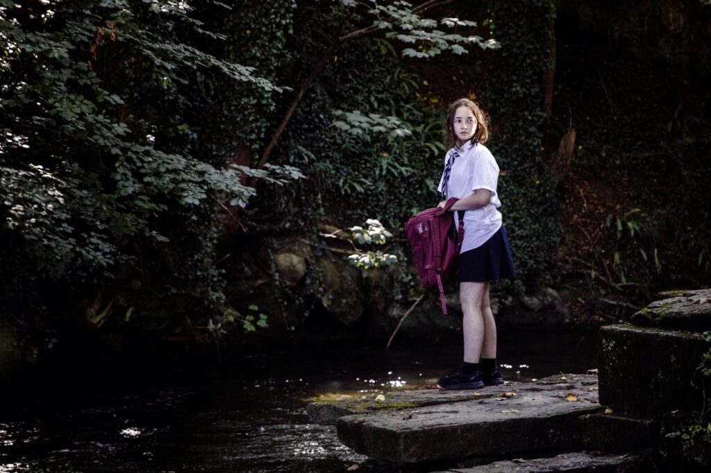 A girl in school uniform holding a backpack, standing on stone slabs over a stream in dense woodland, looking back over her shoulder