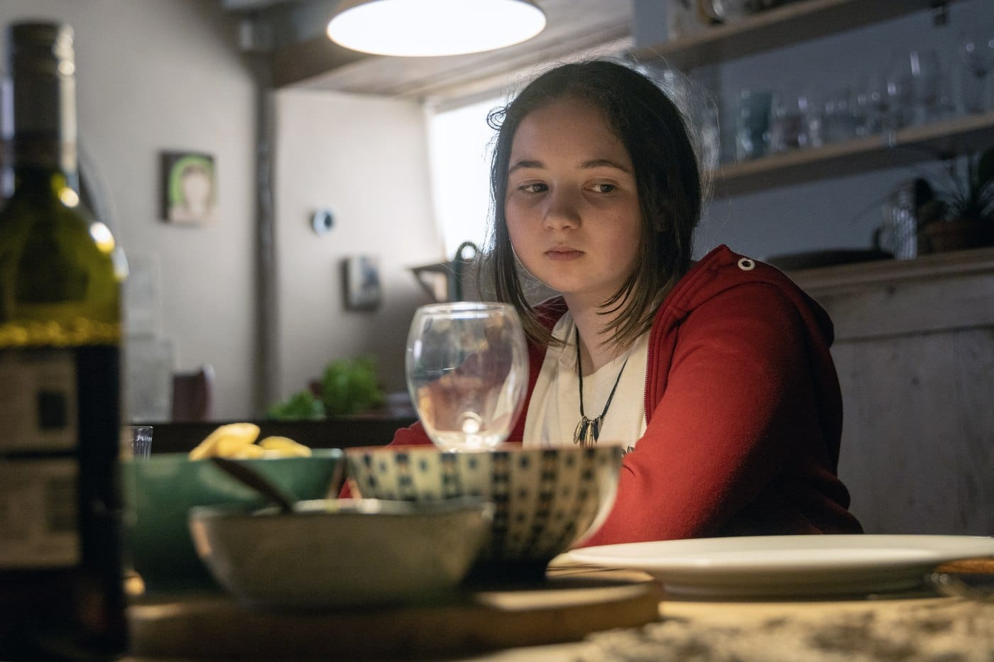 A girl in a red hooded top sitting at a laid kitchen table behind a wine glass and serving bowls, looking off camera