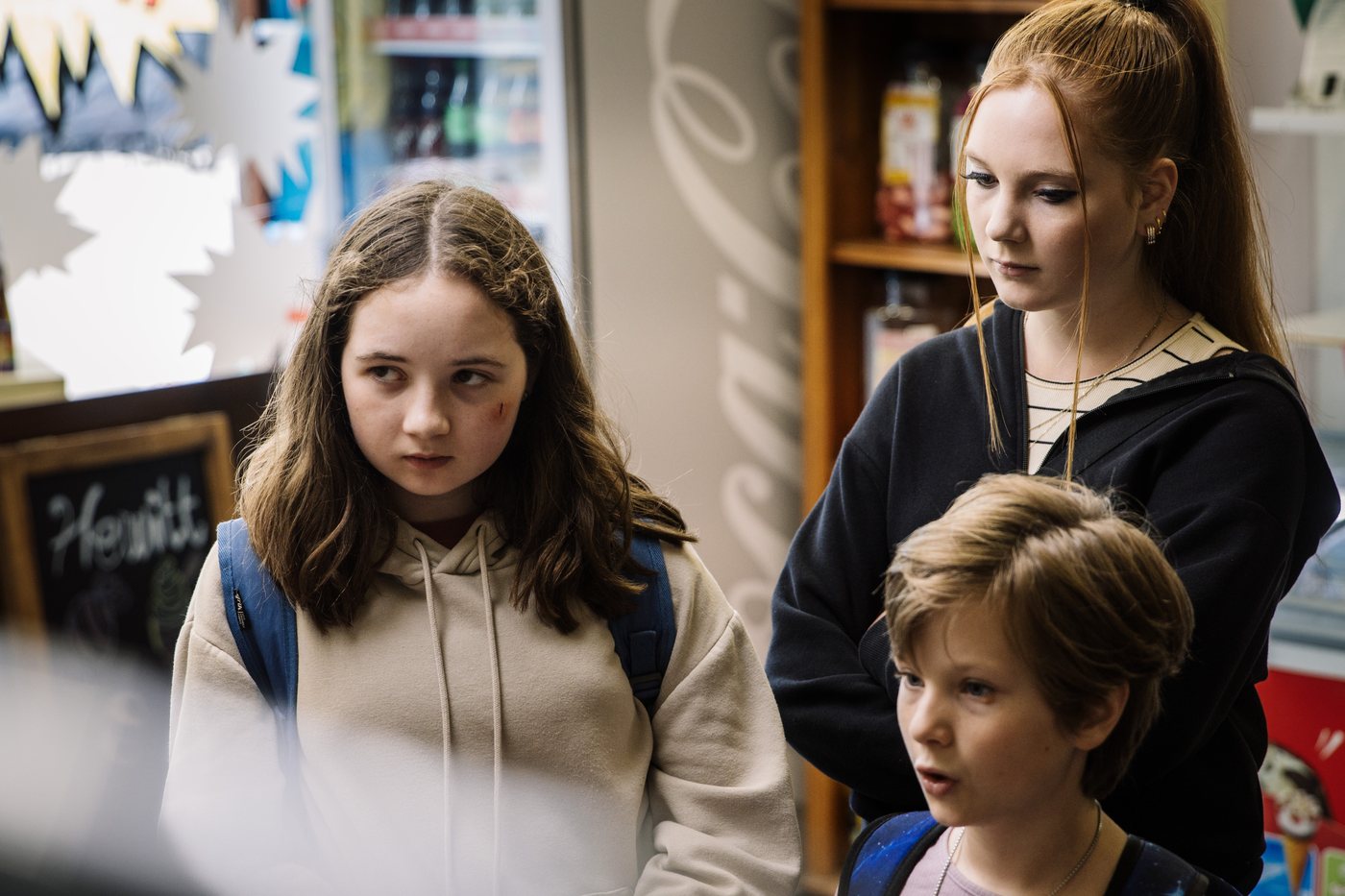 Three children standing in a village shop, the eldest girl with a graze on her cheek looking sideways