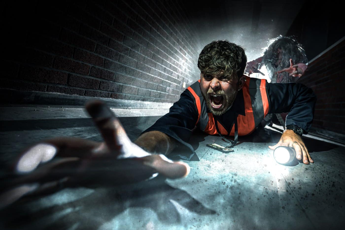 A man in a hi-vis vest sprawled on the floor of a dark corridor, shouting, with a hand reaching towards him and a figure behind