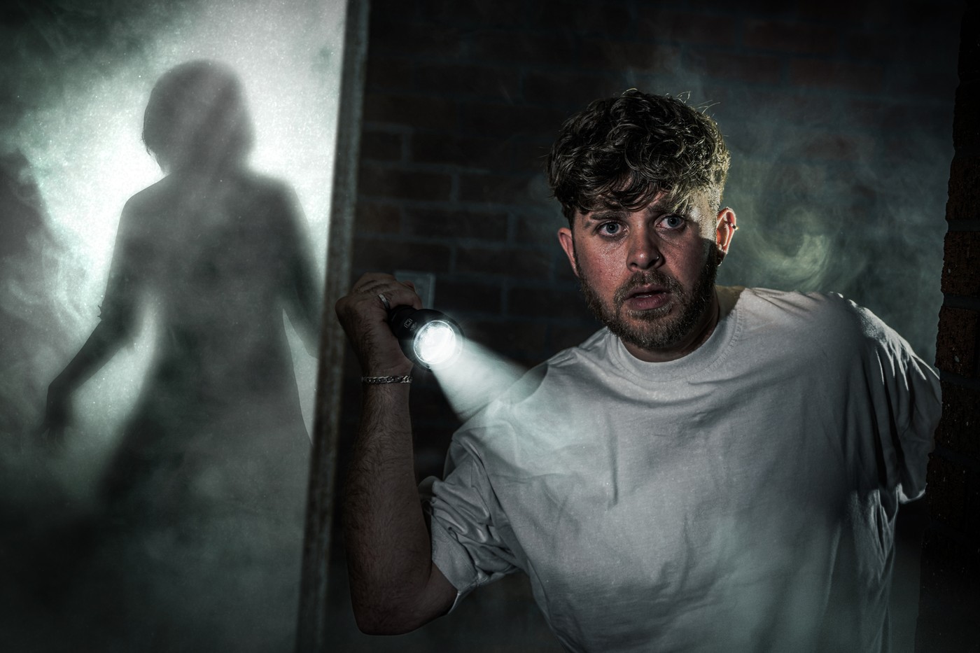 A man holding a torch in a dark brick corridor, with a figure silhouetted in smoke behind him