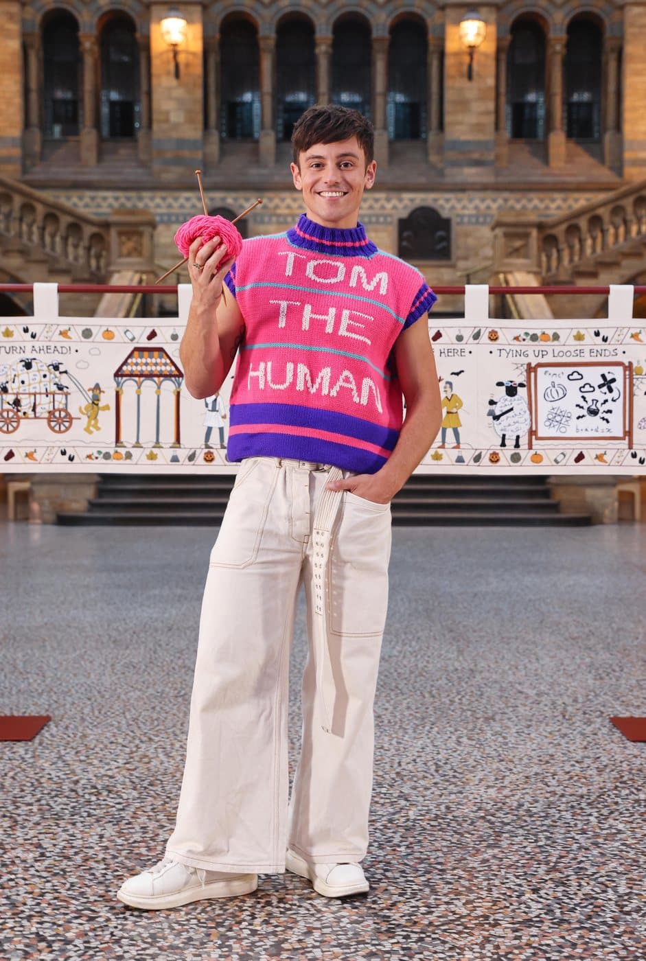 Tom Daley in a pink and purple knitted sweater vest reading Tom the Human, holding wool and knitting needles in front of the Baa-Ewe Tapestry at the Natural History Museum