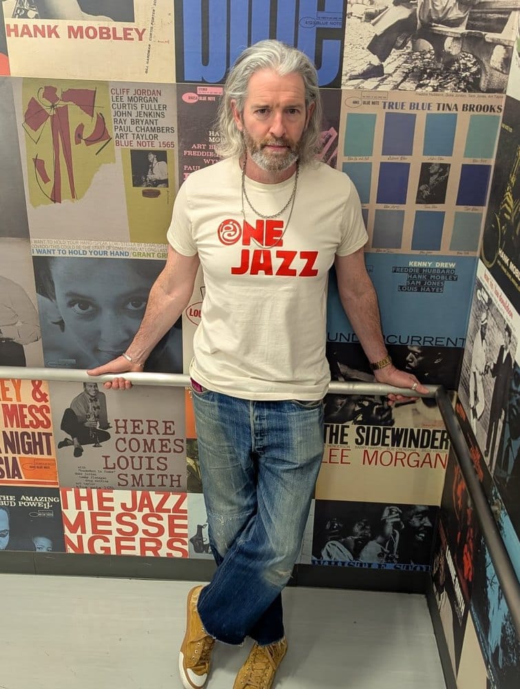 Tony Higgins, The Jazz Dad, in a cream One Jazz t-shirt standing against a wall papered with Blue Note album sleeves