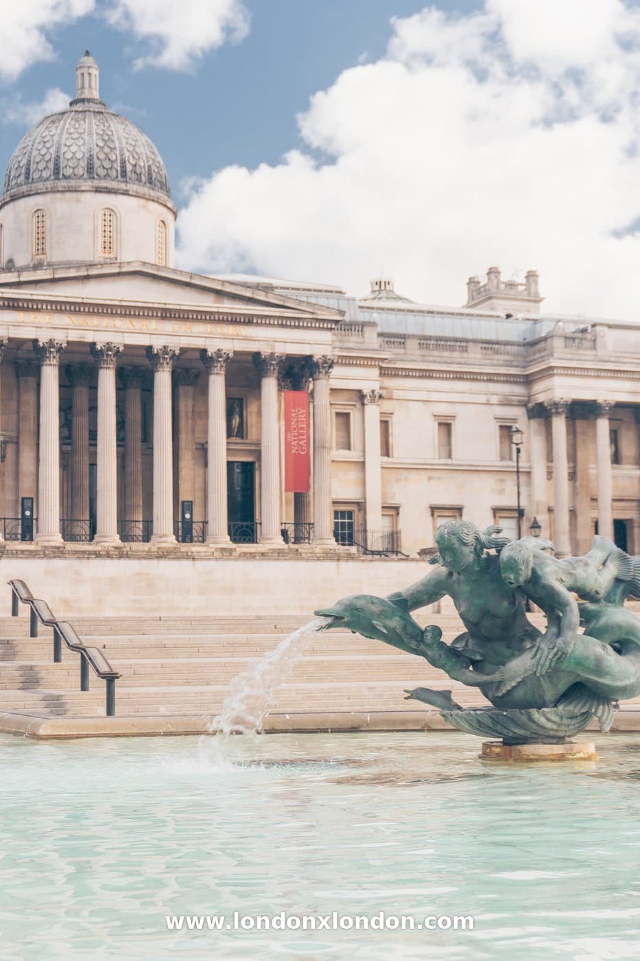 Water arcing from a bronze dolphin into a Trafalgar Square fountain basin with the National Gallery dome above
