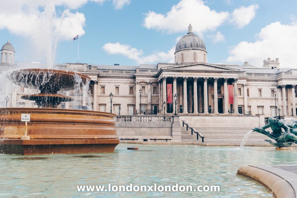 The tiered stone basin and water jet of a Trafalgar Square fountain with the National Gallery behind