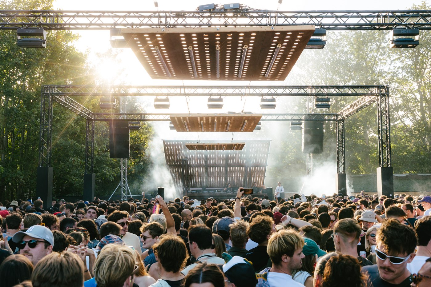 A daytime festival crowd in front of a stage rigged with hanging light panels, sun breaking through the trees