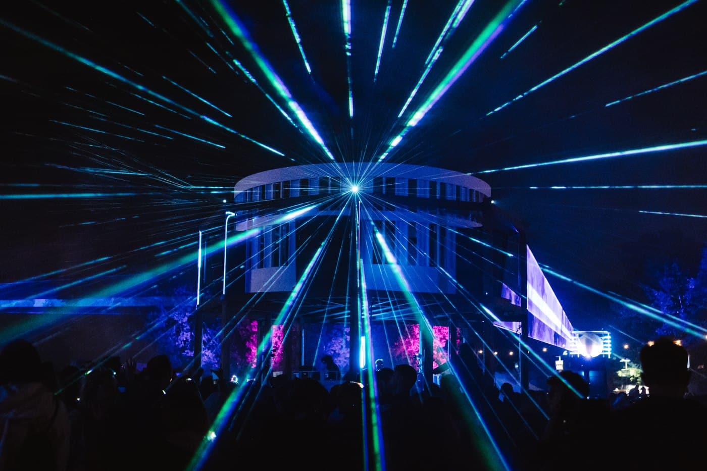 Green and blue lasers bursting outward from a circular building over a Waterworks crowd at night
